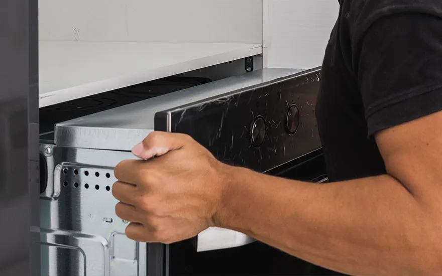 Monteur schuift een metalen inbouwoven in een keukenkast, houdt het toestel stevig vast aan de zijkant met gespannen arm.