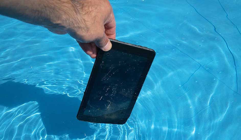 Een hand houdt een zwarte tablet deels onder in helder blauw zwembadwater; op het scherm zitten druppels en rimpelingen vormen kringen.