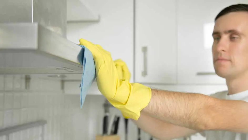 Man met gele handschoen en blauwe doek maakt een zilverkleurige afzuigkap schoon in een witte keuken.