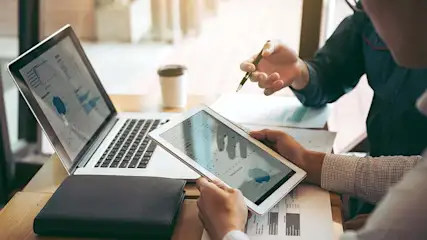 Een laptop en een tablet met grafieken staan op een bureau met documenten. Twee personen bespreken de data met een pen.