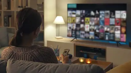 A woman sits on a sofa, remote in hand, watching a TV displaying many movies. A lamp and shelves are in the background.