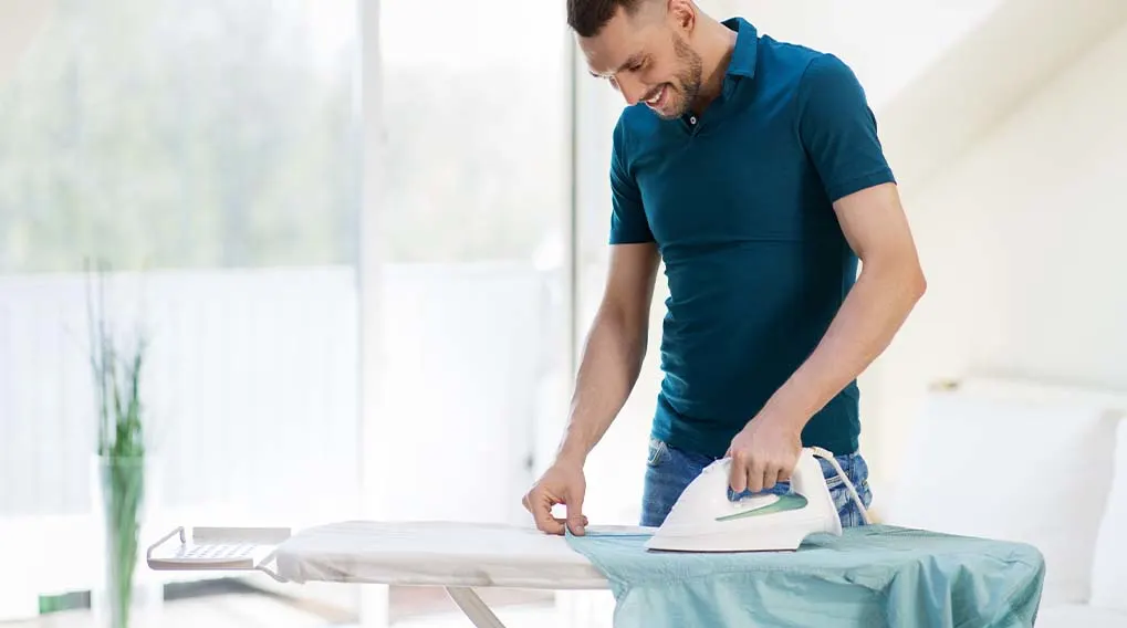 Een man strijkt een blauw overhemd op een strijkplank in een lichte kamer. Op de achtergrond zijn een raam en een vaas met planten zichtbaar.