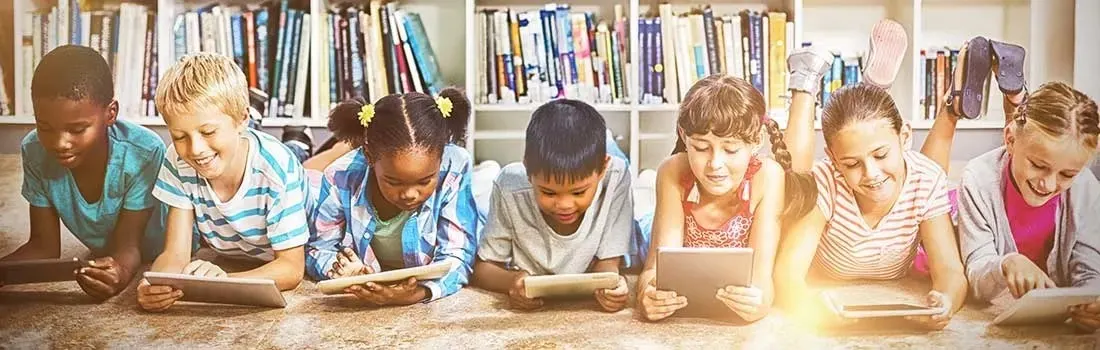 Six children are lying on the floor in a library using tablets. There are many books in the background. No brands are visible.