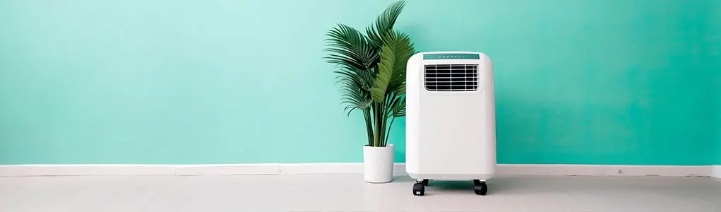 A white portable air conditioner stands next to a potted plant on a light floor against a teal wall.