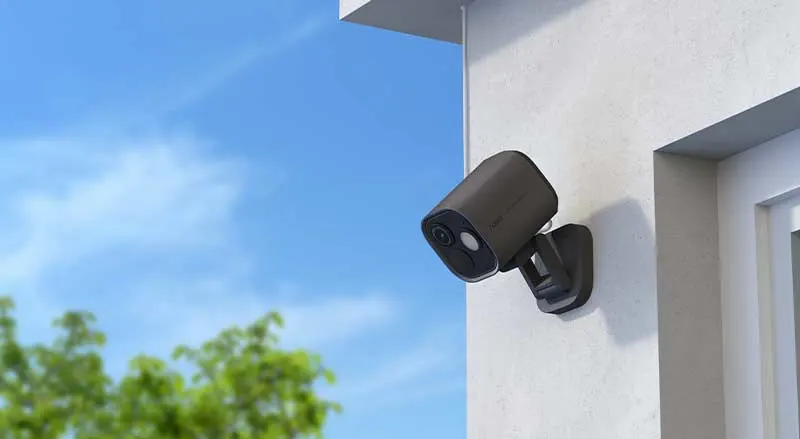 Een zwarte buitencamera hangt aan een moderne gevel en is gericht op buiten, tegen een heldere blauwe lucht.