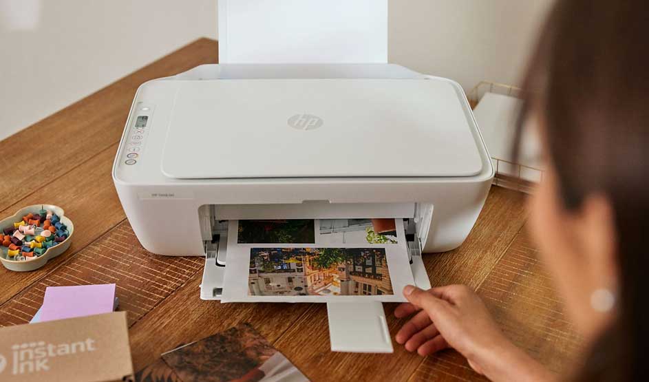 Een vrouw pakt een foto uit een witte HP printer op een houten tafel, met naast de printer een bakje en HP Instant Ink doosje.