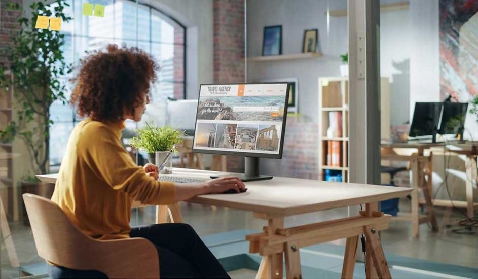 Woman in yellow jumper working at a desk, viewing travel photos on a monitor in a bright, modern industrial office.