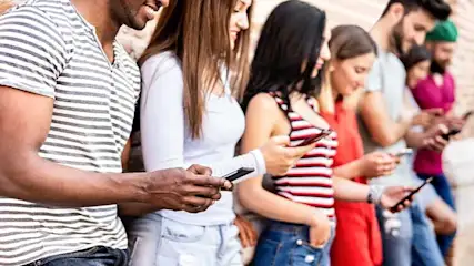 A group stands against a brick wall, each looking at a mobile phone held in their hands. They wear casual clothes.