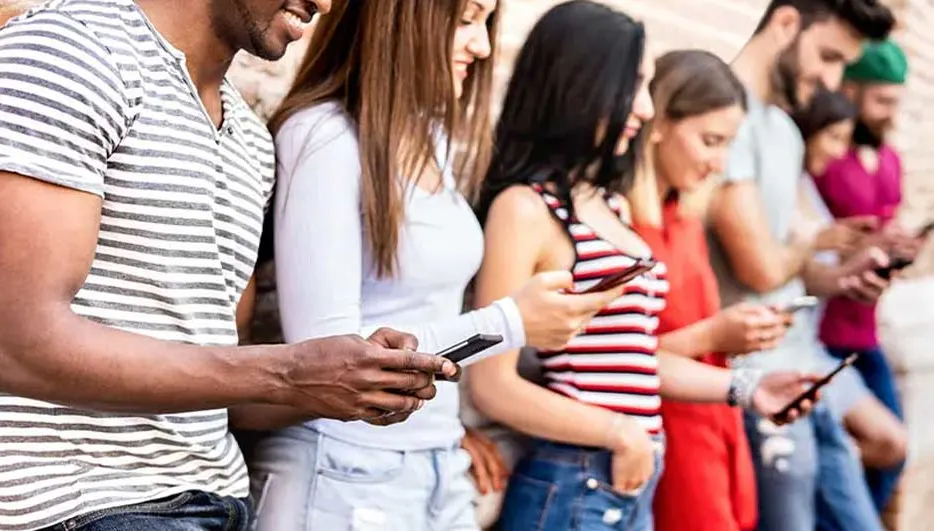 Een groep mensen staat buiten, leunend tegen een muur, en kijkt naar hun smartphones. Ze dragen casual kleding.