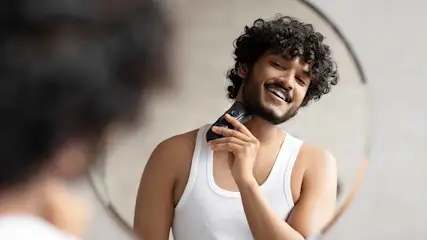 Een man lacht en gebruikt een elektrische scheerapparaat tegen zijn hals voor een spiegel. Hij draagt een wit hemd.