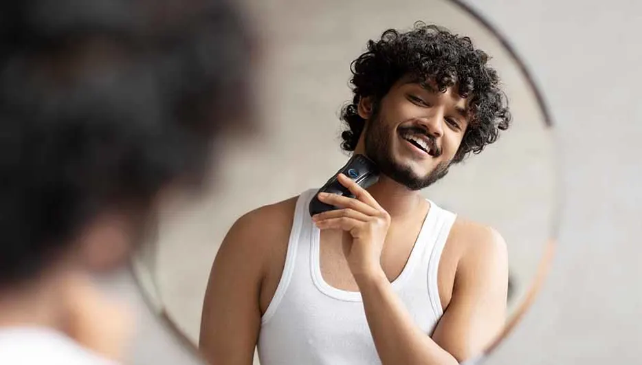 Een man lacht en gebruikt een elektrische scheerapparaat tegen zijn hals voor een spiegel. Hij draagt een wit hemd.