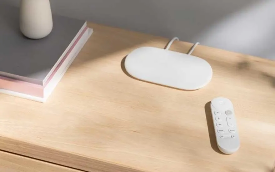 Op een houten kast liggen een minimalistische witte afstandsbediening naast een ovaal streamingapparaat en een stapel designboeken.