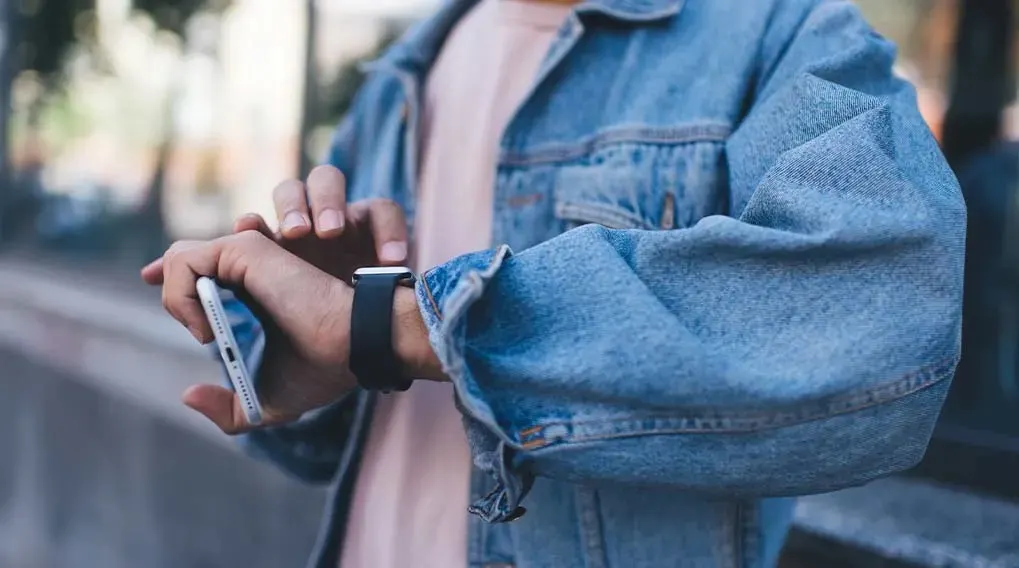 A person in a denim jacket checks their smartwatch while holding a smartphone, walking beside a low street wall.