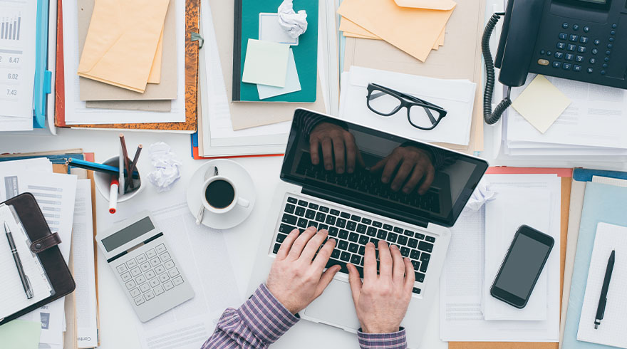 Een bureau met een laptop in het midden, omringd door papieren, een telefoon, een rekenmachine en kantoorbenodigdheden.