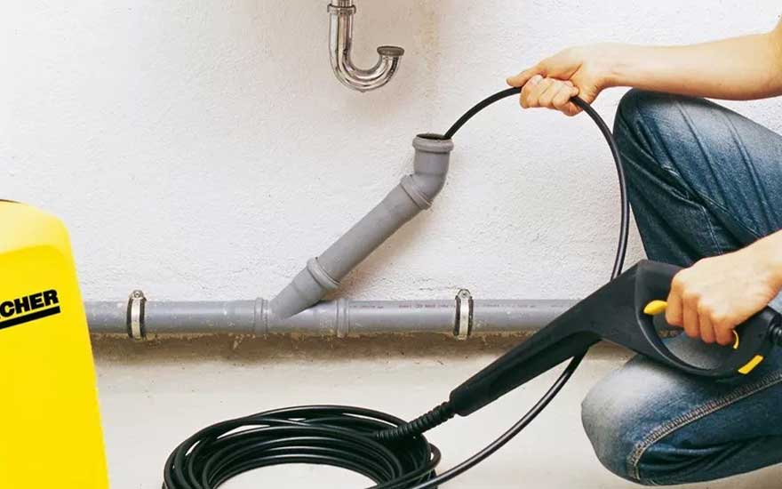 Een persoon gebruikt een Kärcher hogedrukreiniger om een grijze afvoerpijp onder een wasbak schoon te maken. De gele hogedrukreiniger staat links in beeld. De gebruiker houdt het zwarte spuitpistool vast en richt het op de pijp tegen een witte muur.