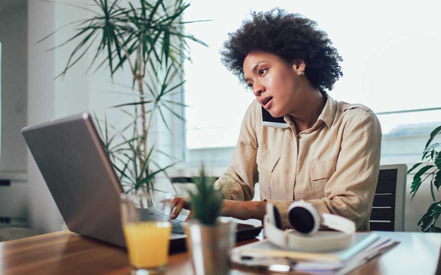 Woman talking on the phone while working on her laptop at a wooden desk with plants.