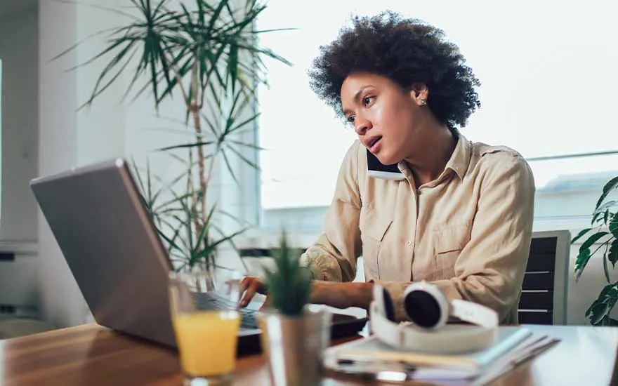 Woman talking on the phone while working on her laptop at a wooden desk with plants.