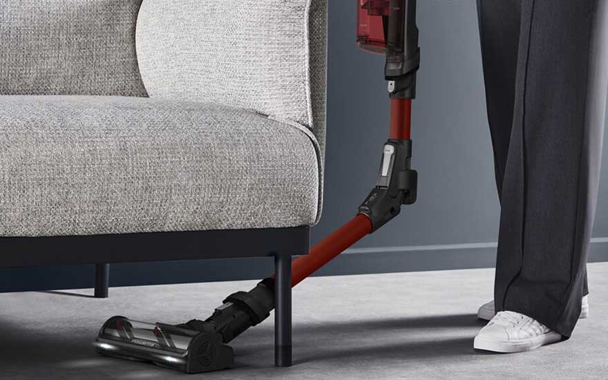 Een flexibele steelstofzuiger buigt onder een grijze fauteuil en reinigt moeiteloos de vloer, terwijl iemand ernaast staat.