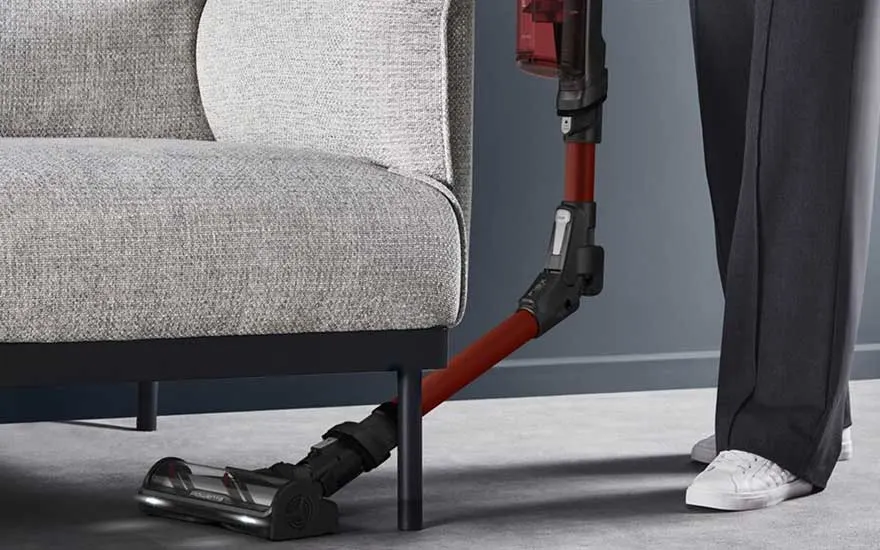 A flexible cordless vacuum bends under a grey armchair, cleaning the floor with ease while a person stands beside it.
