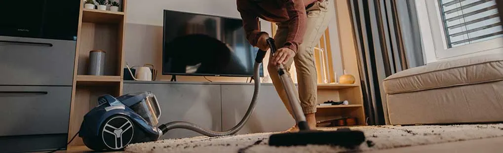 A person carefully vacuums a pale rug in a modern, cosy living room with TV unit, large sofa and warm daylight streaming inside.