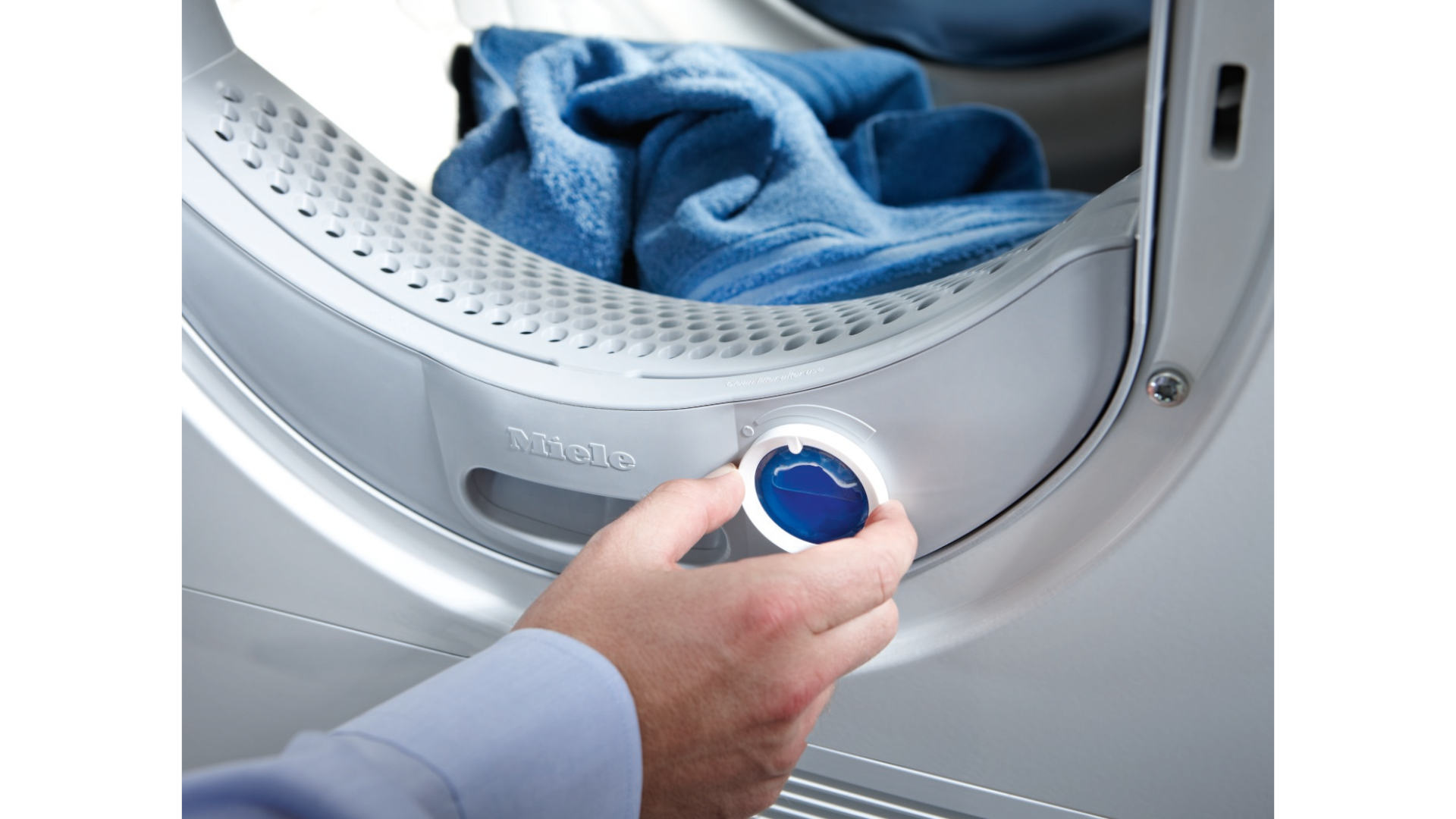 Een hand draait aan een witte knop op een grijze Miele wasdroger met blauwe handdoeken erin.