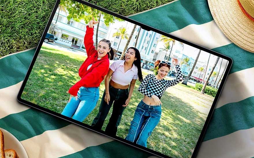 Op een picknickkleed ligt een tablet waarop drie lachende vrienden buiten dansen in een zonnig park vol palmbomen en moderne gebouwen.