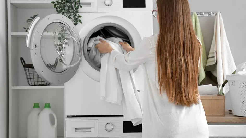 Een vrouw laadt wit wasgoed in een witte wasmachine in een lichte wasruimte met rekken en een mand.