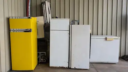 Gele en witte koelkasten en een diepvriezer staan tegen een wand van verticaal geprofileerde metalen platen.