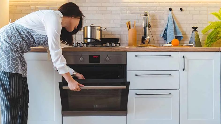 Een vrouw opent de oven in een keuken met witte kasten en houten aanrechtblad. Een pan staat op het fornuis.