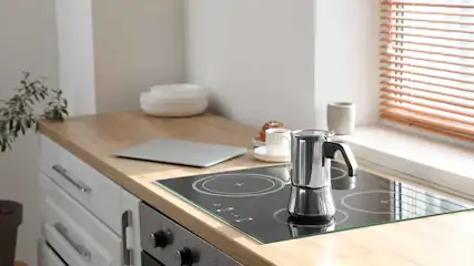 Een keuken met een kookplaat waarop een mokkapot staat, een laptop en een kopje op het aanrecht.