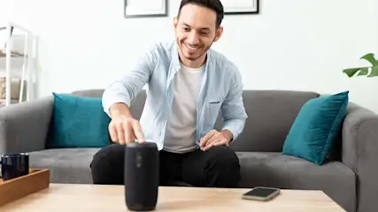 Man zit op een grijze bank met blauwe kussens en bedient een zwarte speaker op een houten tafel. Een telefoon ligt naast hem.