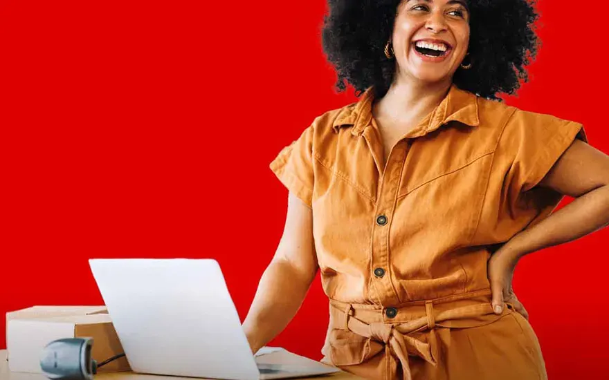 Vrouw lacht bij een laptop met pakket en scanner op tafel, tegen een rode achtergrond.