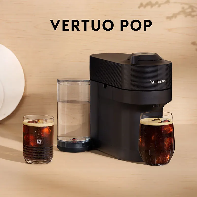 Een zwarte Nespresso machine met twee glazen met ijskoffie en een waterreservoir op een beige achtergrond.