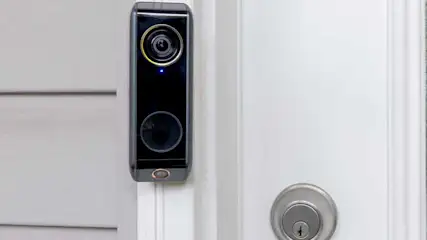 Een zwarte slimme deurbel met een camera aan een witte deur, naast een zilverkleurig slot.