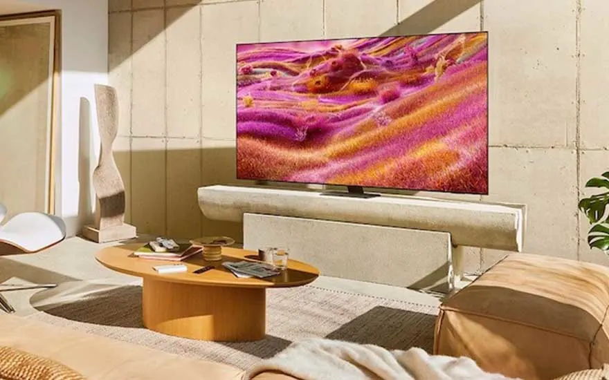 Moderne woonkamer met grote TV die levendig paars-oranje landschap toont, beige bank, houten salontafel en warme zonlichtinval.