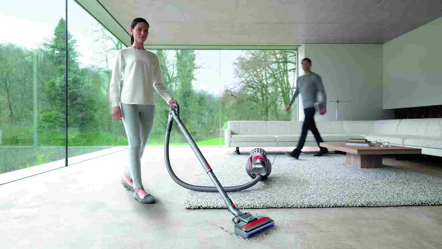 Vrouw stofzuigt een grijze vloerkleed met een rode stofzuiger in een kamer met een witte bank en een man op de achtergrond.