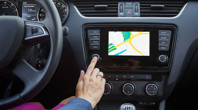 Interieur van een auto met een hand die op een knop drukt, een navigatiescherm en bedieningselementen voor radio en instellingen.