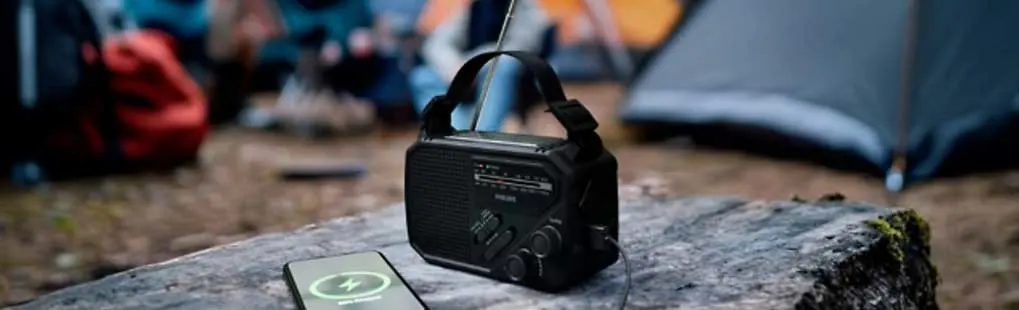 Op een boskamp staat een draagbare zwarte radio op een boomstam, verbonden met een smartphone, terwijl tenten vaag oplichten.