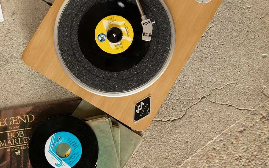 Wooden turntable spinning a black single, surrounded by colourful record sleeves lying casually on a rough concrete floor.
