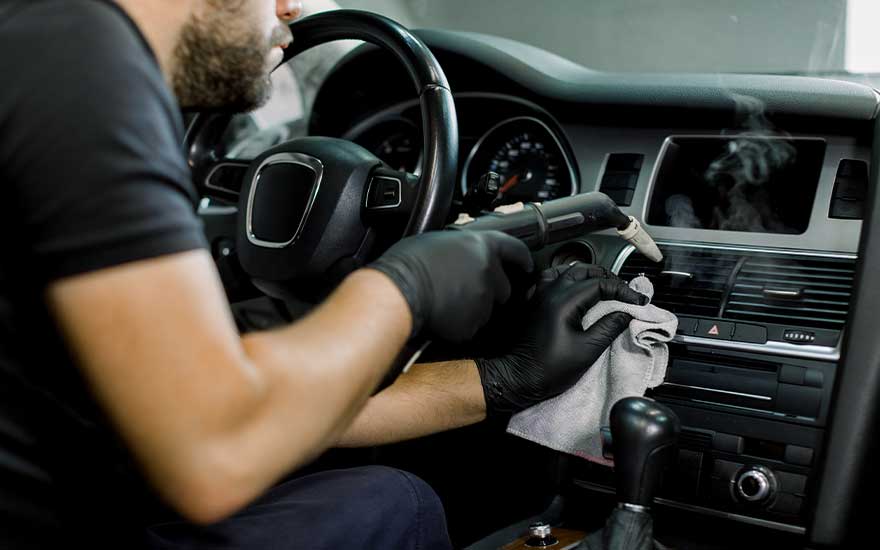 Een man met handschoenen gebruikt een stoomreiniger om het dashboard en ventilatieroosters van zijn auto grondig schoon te maken.