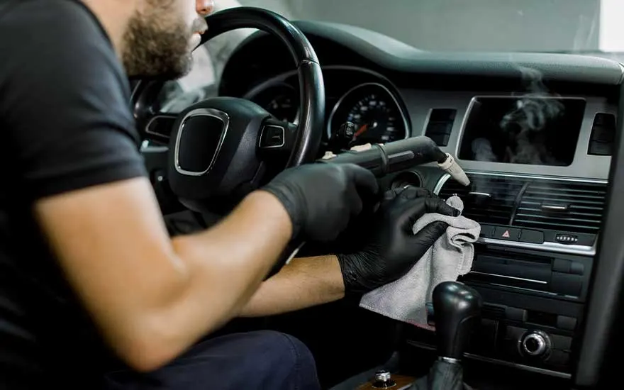 Een man met handschoenen gebruikt een stoomreiniger om het dashboard en ventilatieroosters van zijn auto grondig schoon te maken.