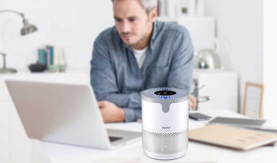 Man working on laptop with white Alpina air purifier on desk in bright home office setting