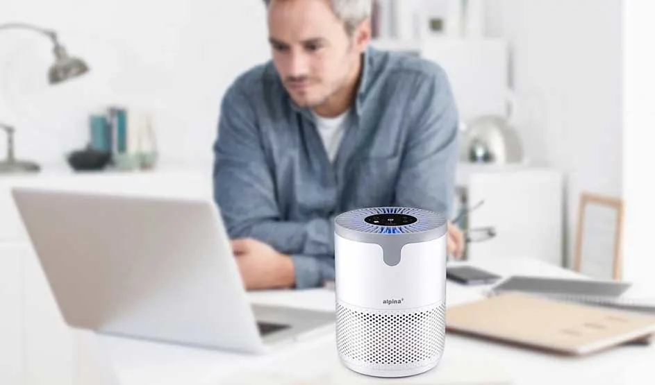 Persoon in blauw overhemd werkt op laptop met een Alpina luchtreiniger op bureau in lichte, moderne werkruimte.