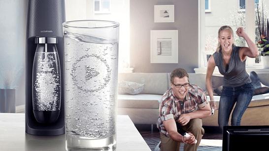 Een bruiswatermaker naast een vol glas water, met een man en een vrouw die een spel spelen op de achtergrond.