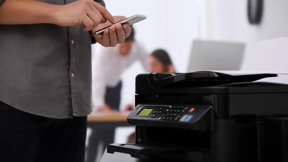 Een persoon gebruikt een telefoon naast een zwarte printer. Achtergrond: een bureau met twee mensen en een computer.