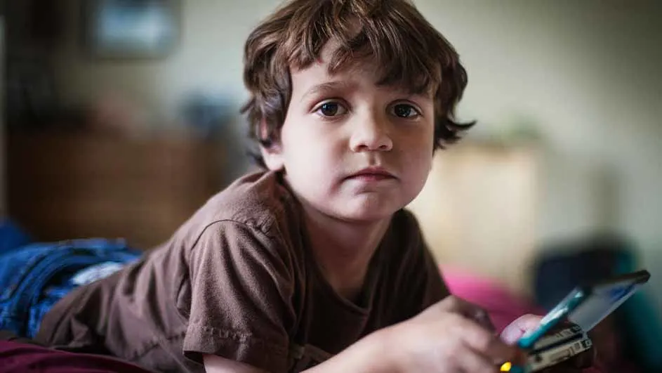 Een kind ligt op een bed met een mobiele telefoon in zijn handen, gekleed in een bruin shirt.