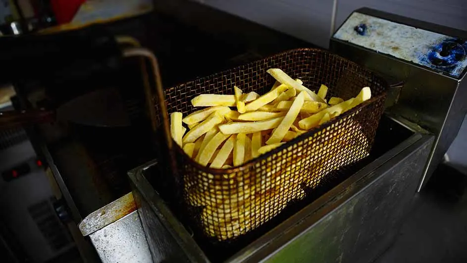 Een metalen mand vol gele, rauwe frieten staat in een friteuse. De friteuse is van metaal en heeft een rechthoekige vorm.