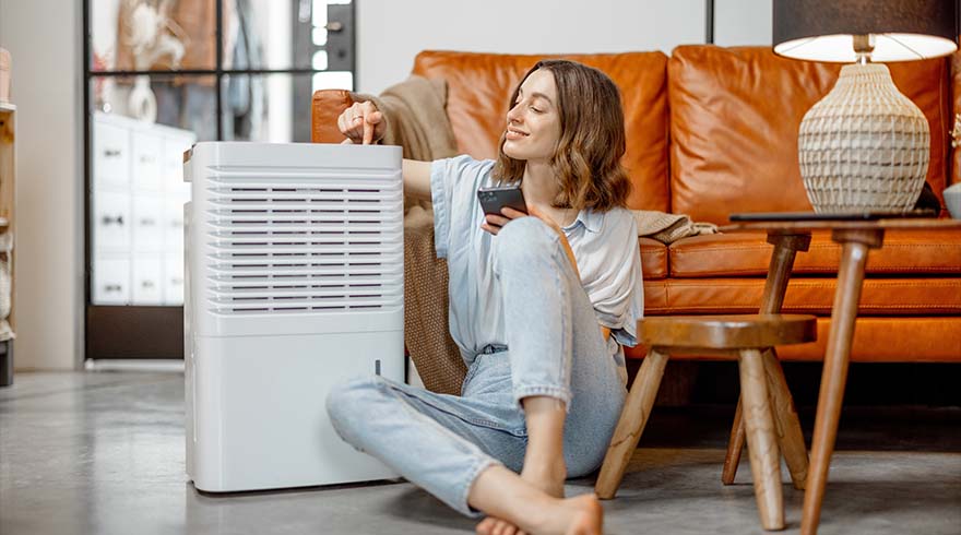 Een vrouw zit op de vloer naast een witte luchtontvochtiger, met een telefoon in haar hand en een oranje bank op de achtergrond.