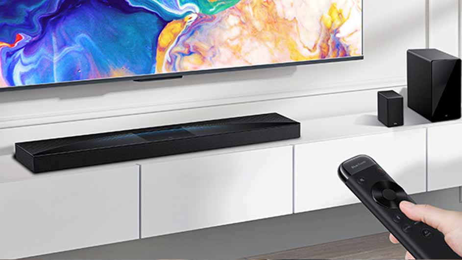 Een zwarte soundbar en luidsprekers staan op een witte kast onder een tv. Een hand houdt een afstandsbediening vast.