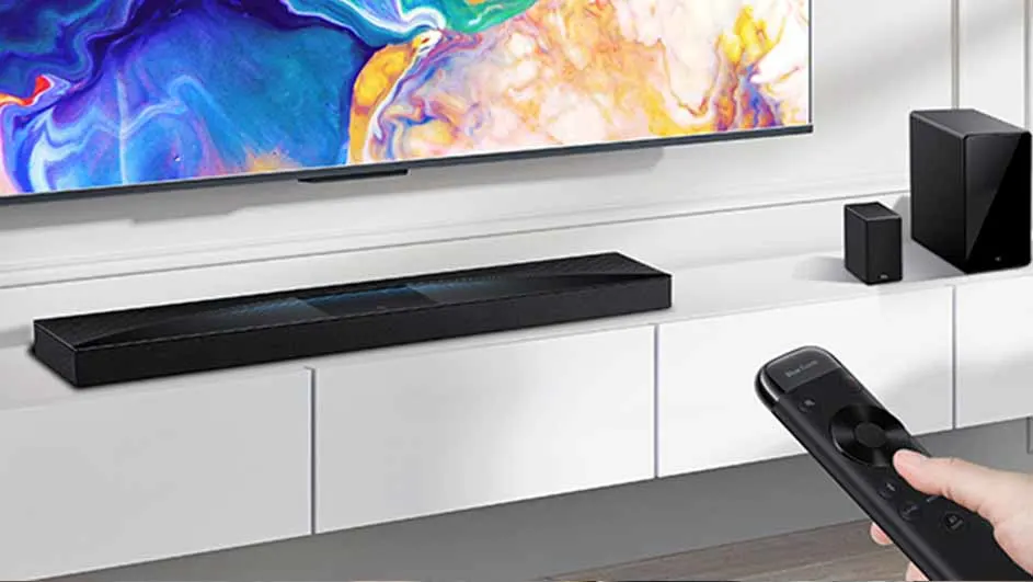 Een zwarte soundbar en luidsprekers staan op een witte kast onder een tv. Een hand houdt een afstandsbediening vast.